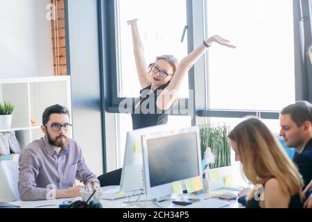 positive woman in glasses with raised arms sitting on the table among serious employees. after good job Stock Photo