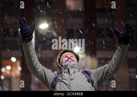Mature Caucasian bearded man in winter jacket standing under snowfall with hands raised to the sky and looking upward Stock Photo
