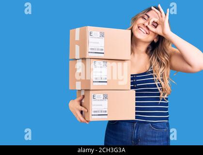 Young caucasian woman holding delivery package smiling happy doing ok sign with hand on eye looking through fingers Stock Photo