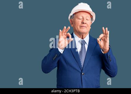 Senior handsome grey-haired man wearing suit and architect hardhat relax and smiling with eyes closed doing meditation gesture with fingers. yoga conc Stock Photo