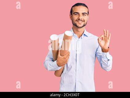 Young handsome man holding paper blueprints doing ok sign with fingers, smiling friendly gesturing excellent symbol Stock Photo