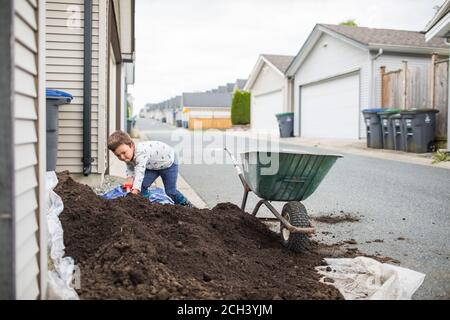 Young boy scooping pile of soil into wheelbarrow in back alley Stock Photo