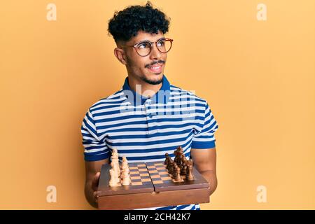 Young arab handsome man holding chess board wearing glasses smiling looking to the side and staring away thinking. Stock Photo