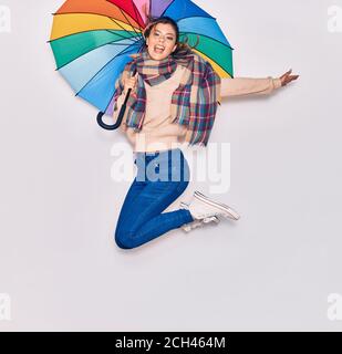 Young beautiful girl wearing winter clothes and scarf smiling happy. Jumping with smile on face holding colorful umbrella over isolated white backgrou Stock Photo