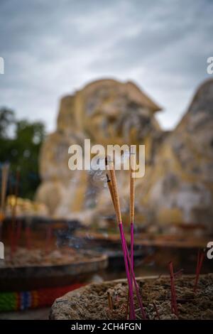 A perspective view of smouldering incense sticks with a large reclining buddha statue in a blurred background. Stock Photo