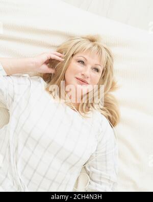 Mature woman with headphones relaxing at home, lying on bed and listening to music. Middle age beautiful blond female smiling and dreaming, self immersion Stock Photo