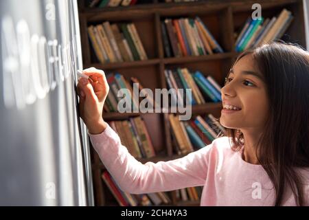 Happy indian latin kid school girl pupil holding chalk writing on blackboard. Stock Photo