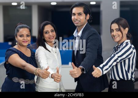 Four Indian office colleagues standing together against office background, showing thumbs up sign of victory, corporate environment. Stock Photo
