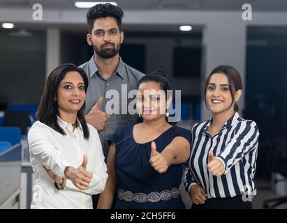 Four Indian office colleagues standing together, showing thumbs up sign of victory, office background, corporate environment. Stock Photo