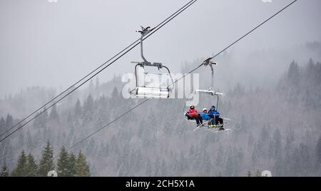 Bukovel, Ukraine - December 09, 2018: family three persons skiers with skis on their feet sitting on cable chairlift carrying up over dark coniferous forest covered with dense fog in winter mountains Stock Photo