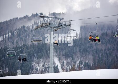 Bukovel, Ukraine - December 09, 2018: skiers and snowboarders sitting carrying high up on cable chairlift in one direction over snow-covered slopes in Carpathians mountains in foggy gray day Stock Photo