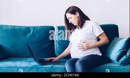Young beautiful pregnant woman working on laptop lying on sofa. Pregnant business woman searching information and online shopping on laptop prepare fo Stock Photo