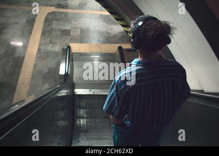 Back view of long haired young man listening to music with headphone standing on escalator inside subway Stock Photo
