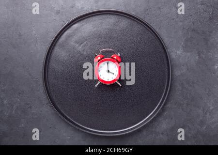 Red alarm clock and empty black ceramic plate on dark stone background. Intermittent fasting concept - Image Stock Photo