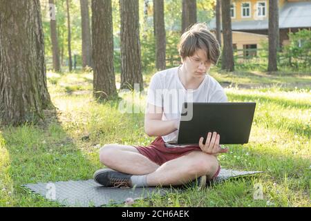 Young man is sitting on the grass in park with his laptop. Distance learning concept. - Image Stock Photo
