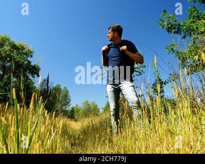 A man tourist with a backpack looks around while standing on a path in summer nature. Stock Photo