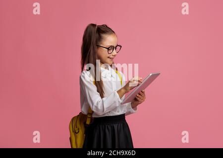 Apps for education. Girl pupil of elementary school in glasses in uniform and with backpack, typing on digital tablet Stock Photo