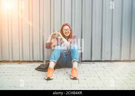 Cheerful Beautiful modern smiling young female teenager in a checkered shirt and jeans with headphones sitting near a wall on the street and making a Stock Photo