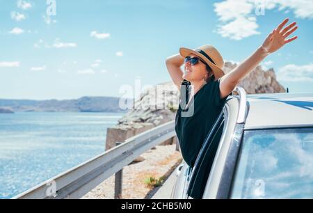 Cheerful Woman portrait enjoying the seaside road trip. Dressed in a black dress, straw hat, and sunglasses she wide opened arms Stock Photo