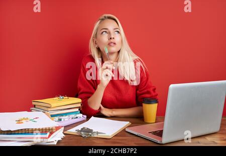 portrait of thoughtful caucasian female at work place, she is contemplation, having too much work, deadlines. sit with laptop isolated over red backgr Stock Photo