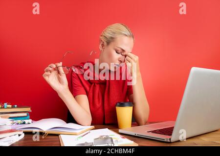 overworked unhappy and frustrated young caucasian woman in stress sitting at office desk, she has a lot of tasks in work, deadline, isolated red backg Stock Photo