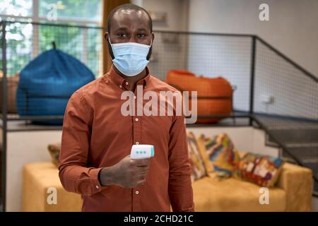Portrait of young african male office worker wearing medical protective mask holding infrared thermometer for checking temperature and looking at Stock Photo