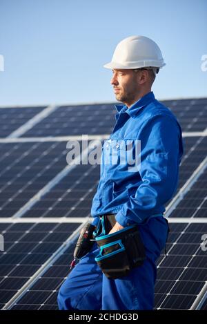 Profile of man engineer in blue suit and protective helmet installing solar photovoltaic panel system using screwdriver. Electrician standing on roof of house. Alternative energy ecological concept. Stock Photo