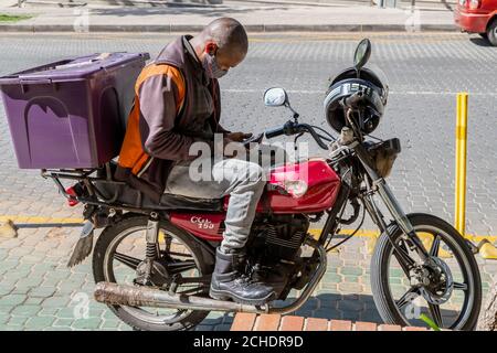 Johannesburg, South Africa, 11th September - 2020: Delivery driver waiting for a delivery order, sitting on motor bike looking at mobile phone. Stock Photo