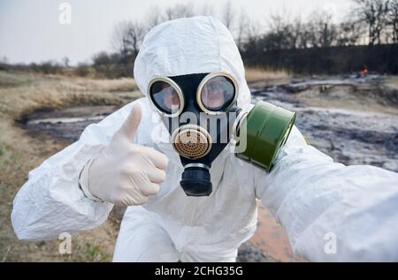 Close-up snapshot of scientist in gas mask and coverall looking right at camera, showing thumbs up, working on contaminated territory, rusted water on background. Concept of polluted nature Stock Photo