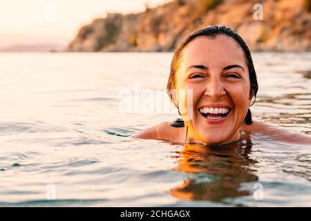 Caucasian woman laughing and showing her white teeth while swimming in sea at sunset