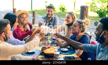 Group of smiling friends wearing protection mask toasting coffee and cappuccino at bar cafe - Young people have breakfast before going to school - The Stock Photo