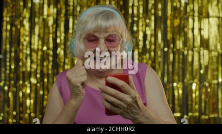 Stylish happy senior old woman in pink blouse holding cell phone, listening to energetic music in headphones, dancing enjoying favorite song. Elderly lady grandma on golden background