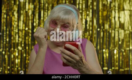 Stylish happy senior old woman in pink blouse holding mobile phone, listening to energetic music in headphones, dancing enjoying favorite song. Elderly lady grandma on golden background