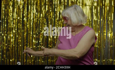 Amazed senior old woman in pink blouse pointing empty place behind her, copy space advertising area on hand, expressing excited surprised emotions. Elderly grandma on golden background Stock Photo