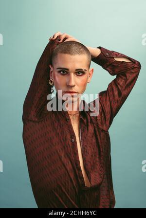 Gay man with makeup wearing earring standing at studio. Transgender male in silk shirt posing over blue background. Stock Photo