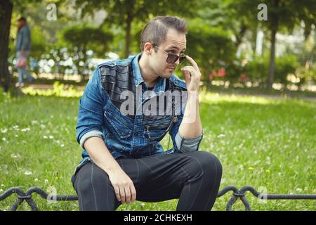 Young white man in sunglasses is sitting in the public park at the daytime. Caucasian male is walking outdoors. Stock Photo