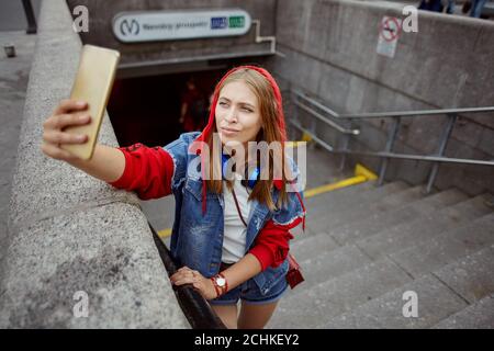 Beautiful young woman in red hoodie is taking selfie using mobile phone near the entrance to the subway station. Stock Photo