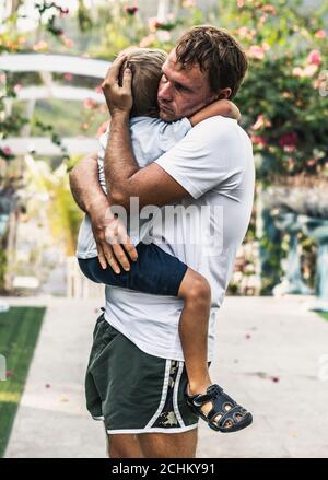 Sad young blond boy crying on fathers hands in nature park outdoor. Man holds son, hugs and comforts. Family love, care and moral support, baby's Stock Photo