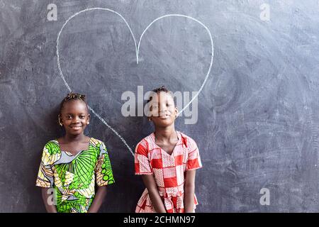 Two Nice Cute African Black Girls Standing in Front of Blackboard with Big Heart Love Symbol Stock Photo