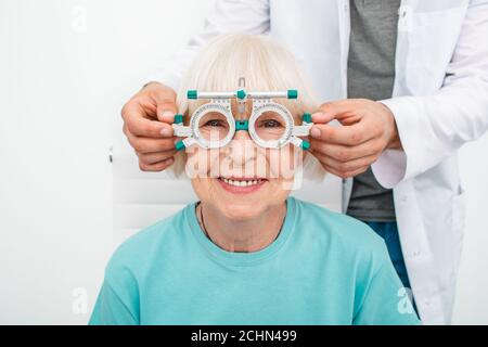 Smiling senior woman wearing optometrist trial frame at ophthalmology clinic. Ophthalmologist helping select glasses for treatment of vision Stock Photo