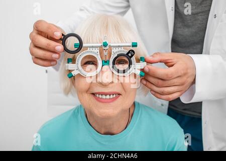 Smiling senior woman wearing optometrist trial frame at ophthalmology clinic. Ophthalmologist helping select glasses for treatment of vision. Eye chec Stock Photo