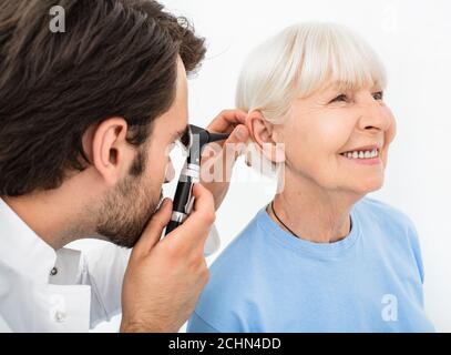 ENT doctor examining senior patient ear, using an otoscope, in doctors office. Elderly smiling woman getting medical ear exam at clinic Stock Photo