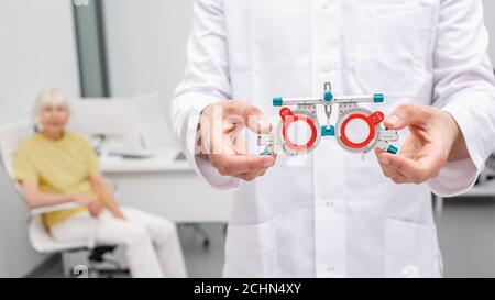 doctor holding an optometric trial frame for eye exam in his hands. Senior woman sitting on background. Concept of checking vision Stock Photo