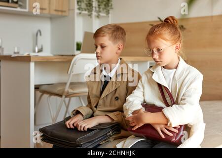 tired kids couple waiting for their taxi in hotel, want to sleep, sad redhead girl and boy sit on bed in coats with bags Stock Photo