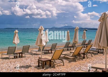 View of empty beach - white deck chairs and umbrellas near sea water, pebbles and golden sand, mountains on the horizon and dark clouds on the sky. Stock Photo