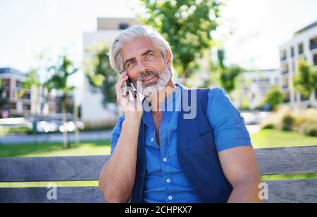 Portrait of mature man sitting outdoors in city, using smartphone. Stock Photo