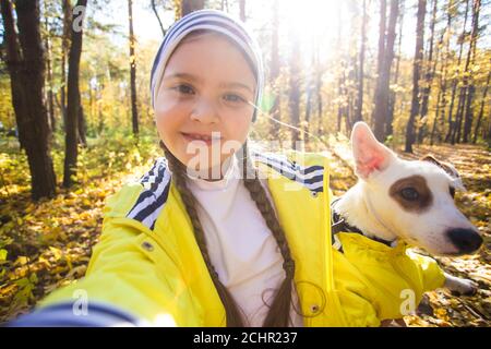 Little girl taking selfie with her dog at autumn park. Child posing with jack russell terrier for a picture on the mobile phone outdoors. Pet and Stock Photo