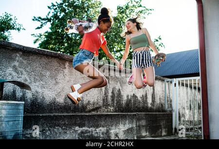 Young teenager girls friends with skateboards outdoors in city, jumping. Stock Photo
