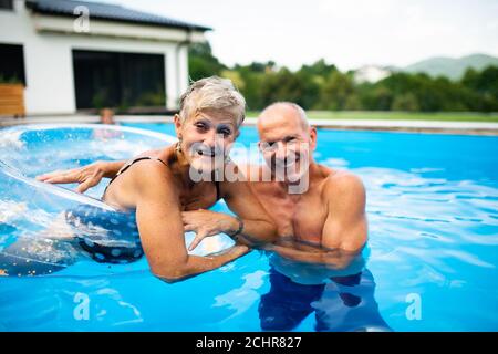 Cheerful senior couple in swimming pool outdoors in backyard, looking at camera. Stock Photo