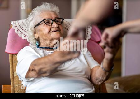 Female home carer supporting old woman to stand up from the armchair at care home Stock Photo